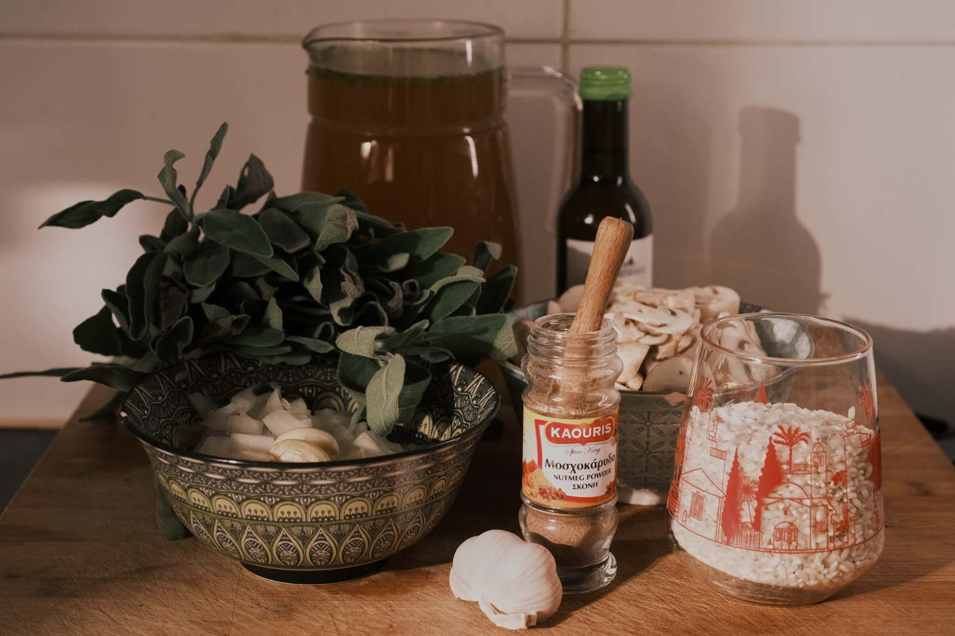 Cooking preparation scene with fresh ingredients laid out on a wooden kitchen surface. Visible are a bowl of chopped onions and whole sage leaves, a jar of honey, a bottle of balsamic vinegar, a glass jar of arborio rice with a red patterned design, a head of garlic, and a jar of ground nutmeg with a red label.