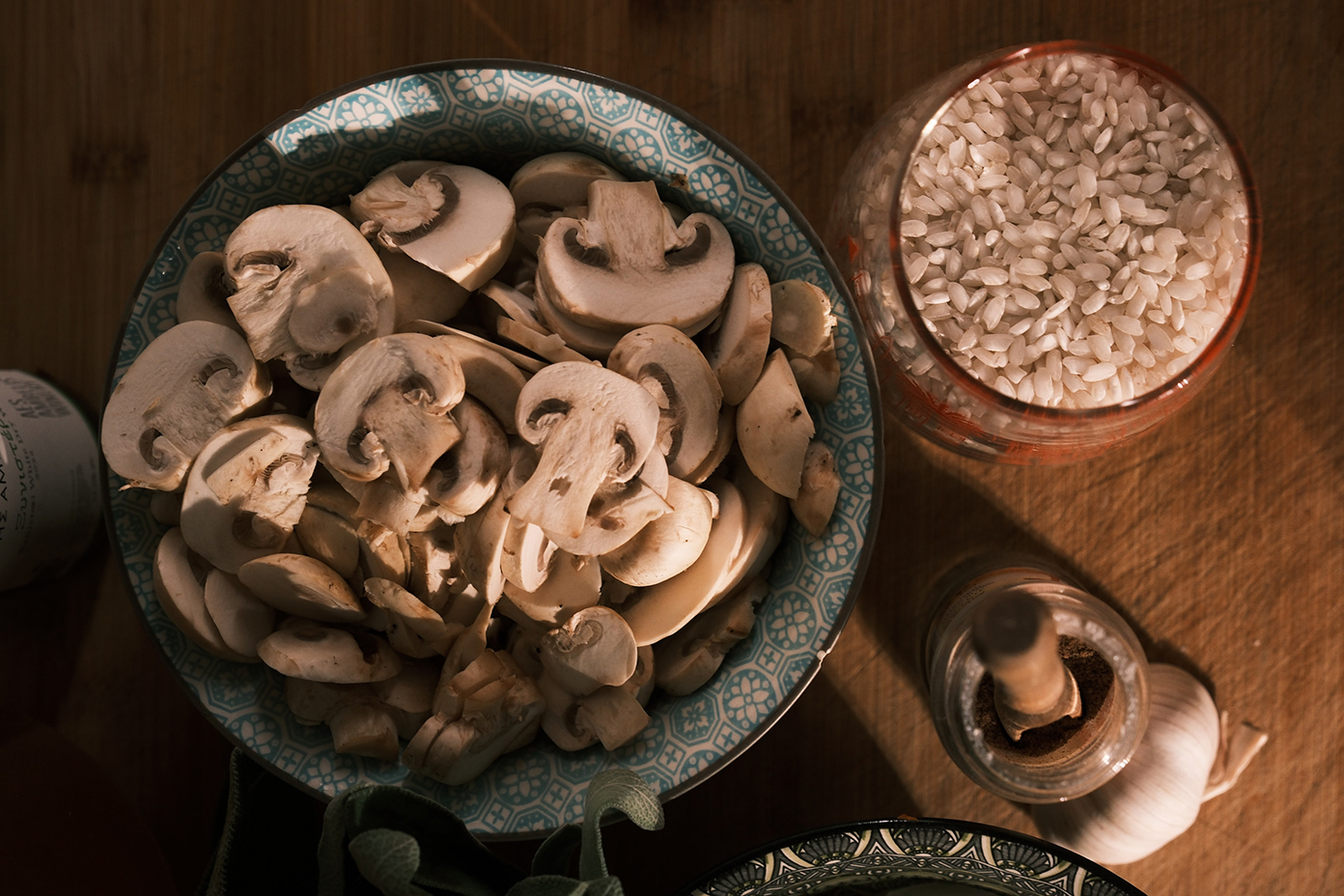 Close-up of a cooking preparation scene showing a detailed view of sliced white mushrooms in a blue patterned ceramic bowl, the glass jar of arborio rice, the jar of ground nutmeg, and the head of garlic, all on a wooden surface bathed in warm light.