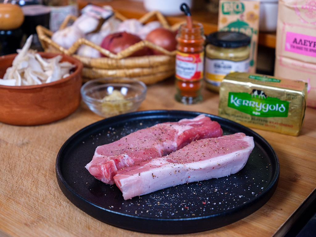 Raw pork chops seasoned with salt and pepper on a black plate, with ingredients like Kerrygold butter, mustard, honey, and various spices in the background on a wooden countertop.