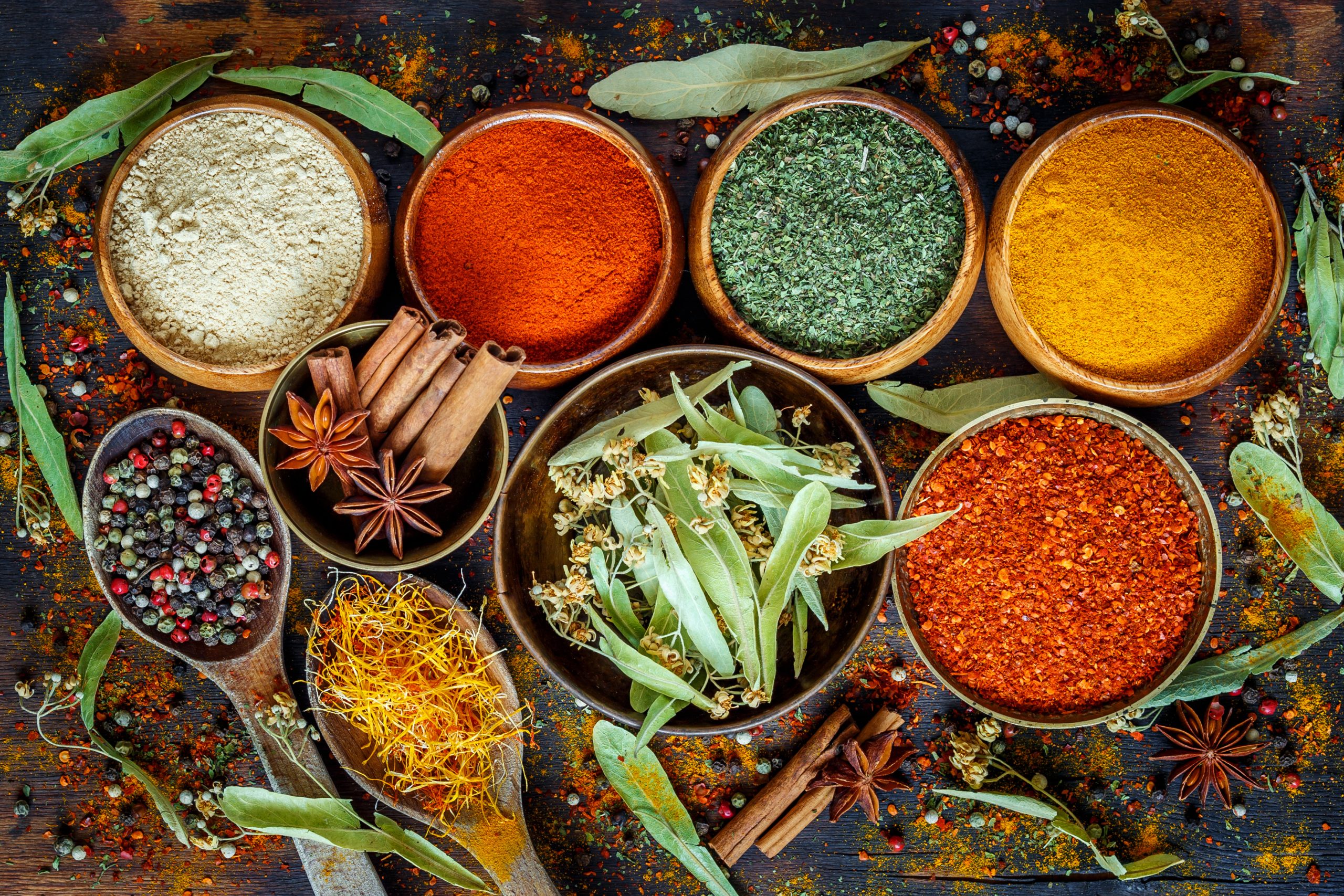 An assortment of spices and herbs displayed in various bowls on a dark wooden surface. The collection includes powdered and whole spices like cinnamon sticks, star anise, and dried bay leaves, with colorful spices such as bright red paprika, golden turmeric, and green dried herbs creating a vibrant contrast. Spilled spices sprinkle the area, adding to the rustic and aromatic atmosphere.