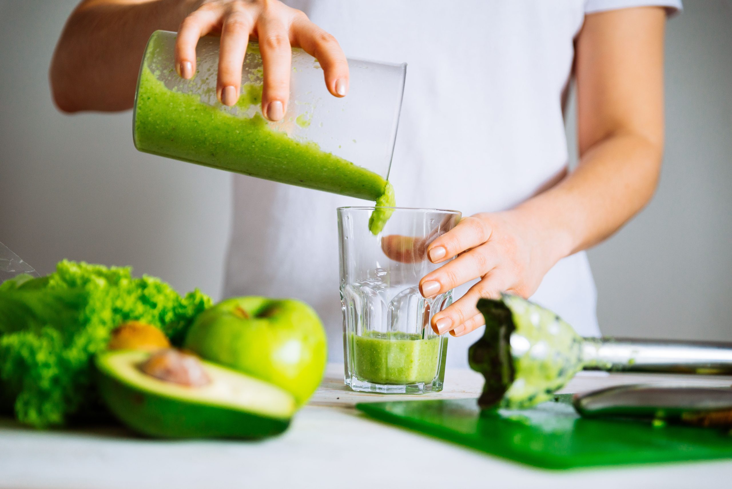 A person pouring a vibrant green smoothie into a clear glass, with fresh green apples, half an avocado, and leafy greens on a cutting board in the background, signifying fresh, healthy ingredients.