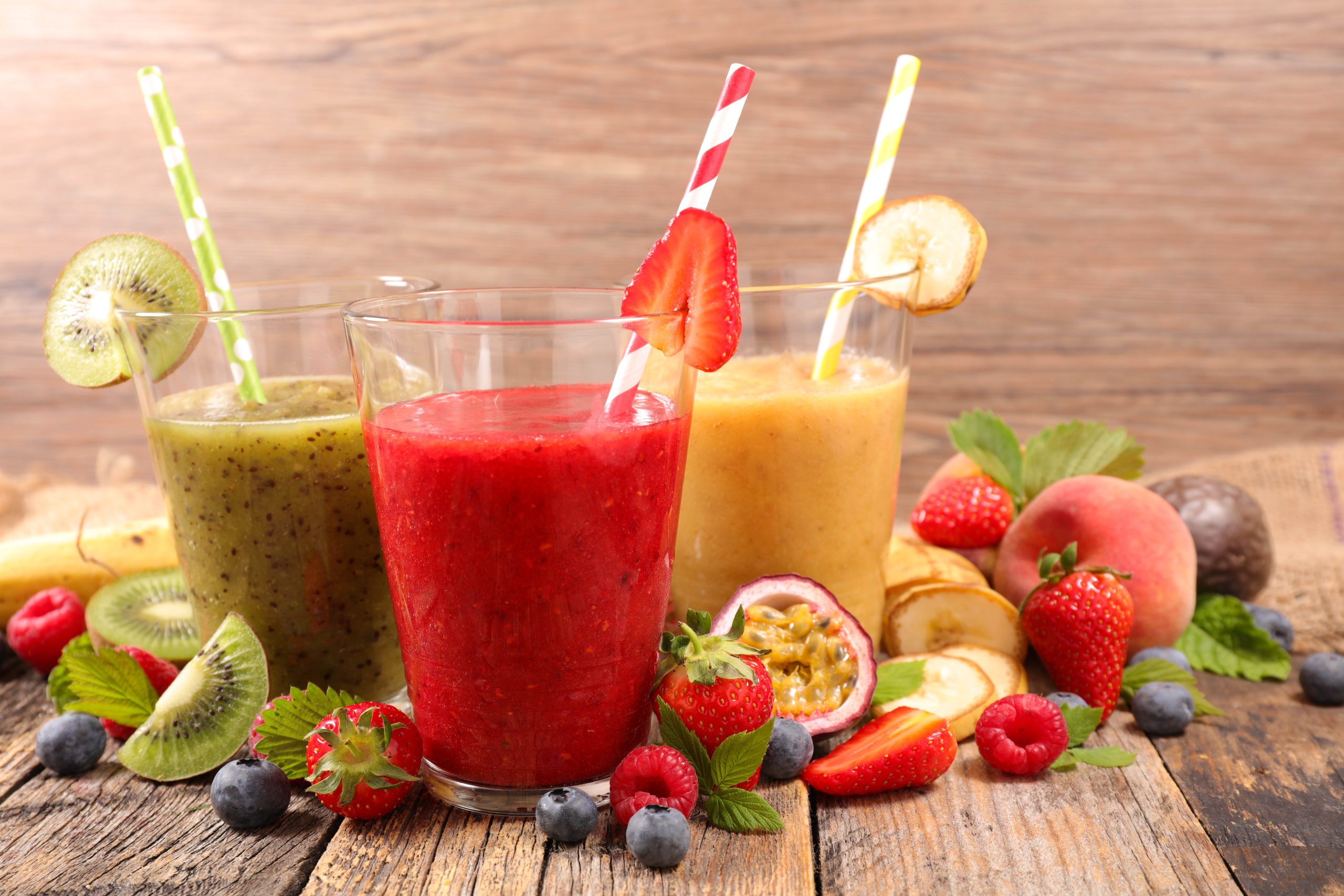 Three colorful smoothies in clear glasses, garnished with kiwi, strawberry, and banana, surrounded by an array of fresh fruits like strawberries, blueberries, raspberries, passion fruit, peach, and mint leaves on a wooden table.