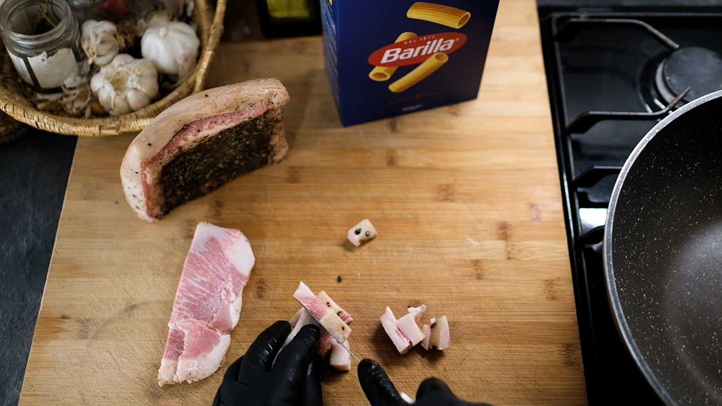  "A kitchen preparation scene with ingredients and cooking utensils on a wooden counter. A piece of raw quanciale is being cut into small pieces, with a person wearing black gloves handling the meat. To the left, a basket with garlic bulbs and a large piece of cured meat with herbs on the rind are visible. In the background, a box of Barilla pasta and a frying pan on a stovetop can be seen, suggesting the cooking of a meal, possibly pasta with bacon."