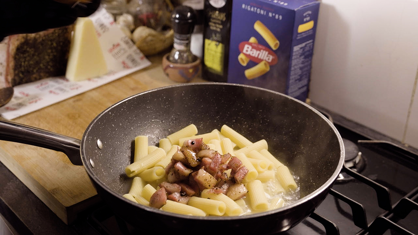  "A frying pan on a stovetop with rigatoni pasta and diced bacon cooking. The pan is in the foreground with steam rising, indicating it's being cooked. In the background, there's a box of Barilla rigatoni pasta, a wedge of cheese, a pepper mill, and a bottle of olive oil, hinting at the ingredients used in this dish. The kitchen scene captures a moment of home cooking, with the focus on the sizzling pan that suggests a cozy, homemade meal is being prepared." 