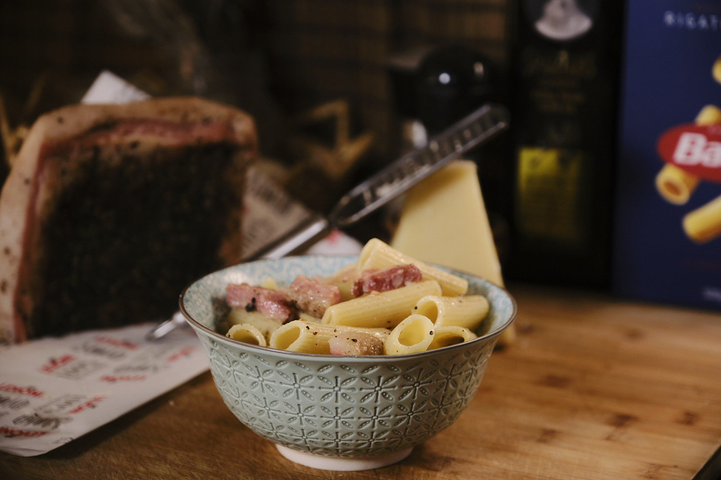 "A finished dish of rigatoni pasta with chunks of pink quanciale served in an ornate blue bowl on a wooden countertop. Behind the bowl, there’s a large wedge of cheese, a bottle of olive oil, and a box of Barilla pasta, suggesting the ingredients used in the recipe. The blurred background includes a cutting board with a large slab of seasoned meat, adding depth and context to the home cooking theme. The image captures a cozy, inviting meal ready to be enjoyed."