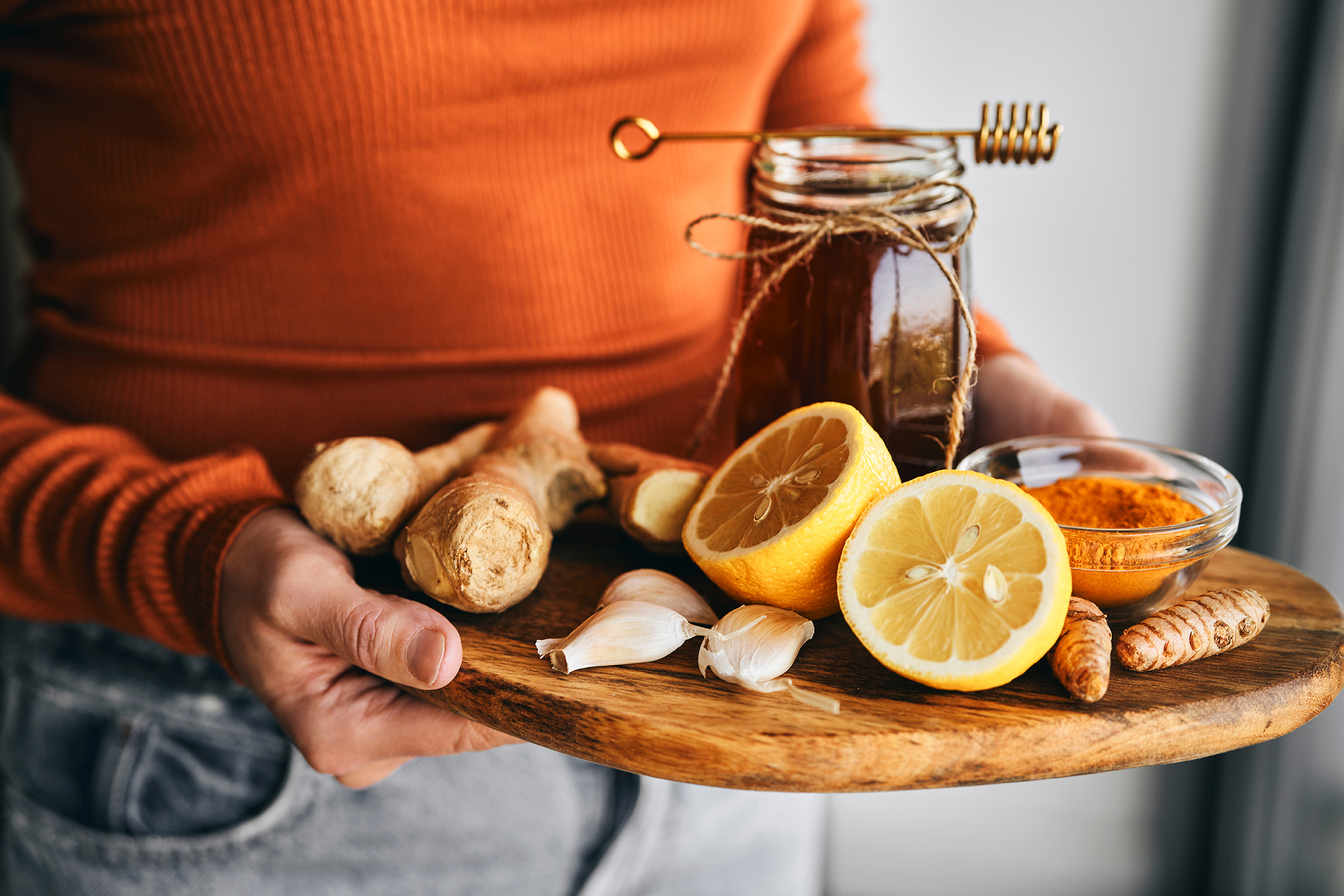 Woman holding a wooden tray with natural cold remedies including ginger root, halved lemons, garlic cloves, a jar of honey with a honey dipper, and a small bowl of turmeric powder."