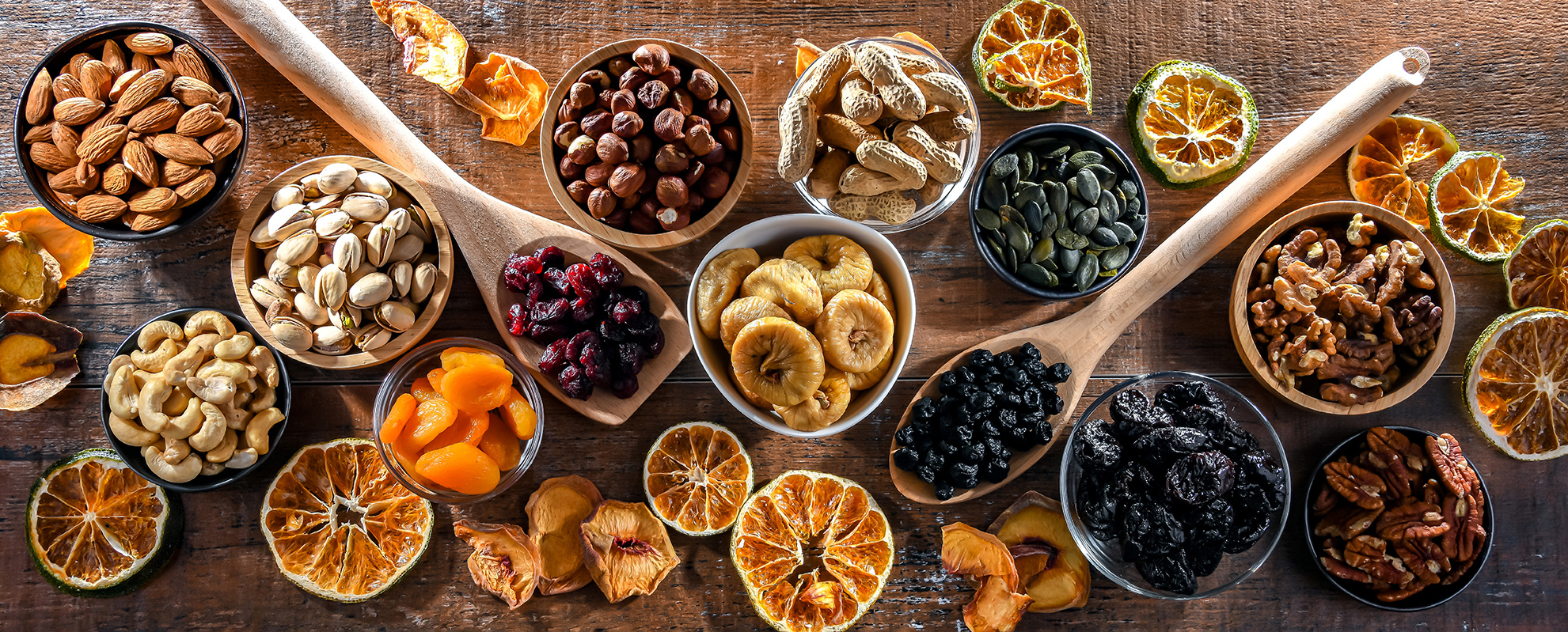  "A panoramic view of various dried fruits and nuts displayed on a wooden surface. The assortment includes almonds, pistachios, hazelnuts, peanuts, pumpkin seeds, cashews, dried cranberries, dried figs, dried apricots, black raisins, and walnuts, along with dried slices of orange. Rolling pins and scattered dried orange slices add to the rustic presentation."