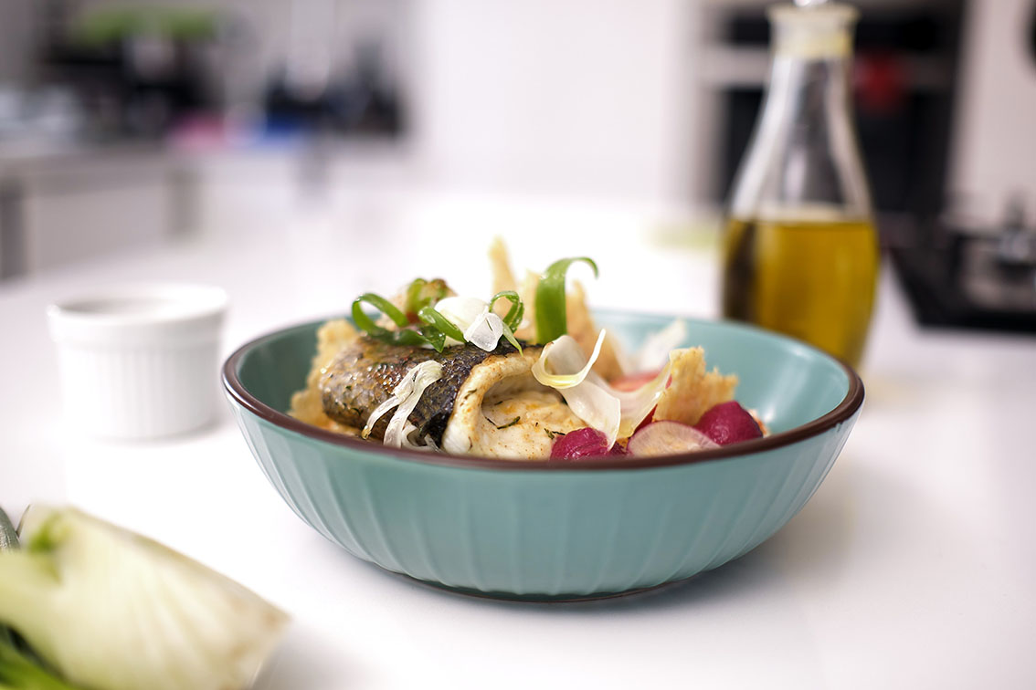Pan-seared fish fillet with sliced radishes and onions, garnished with green onion tops, counter with a bottle of olive oil and a ramekin in the background